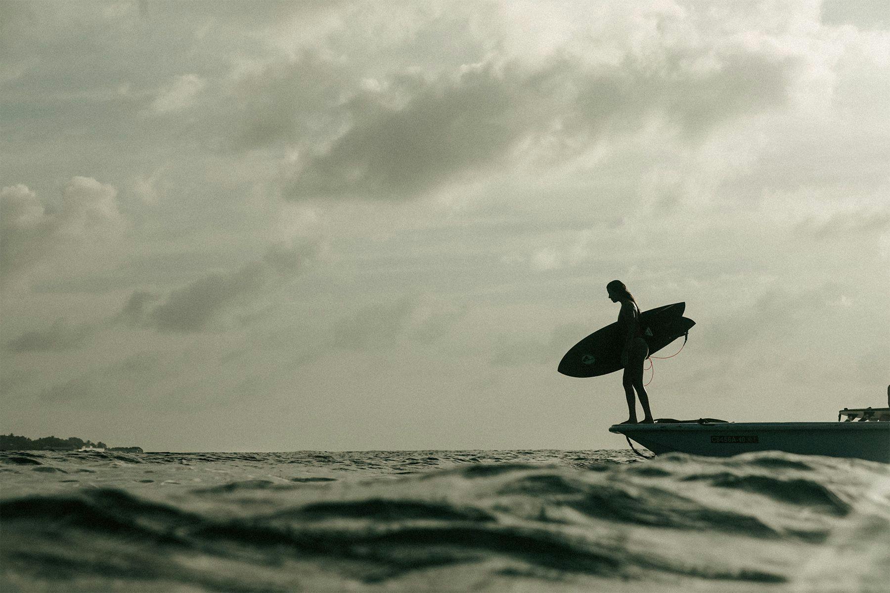 silhouette of a female surfer holding a fish surfboard on the bow of a small boat