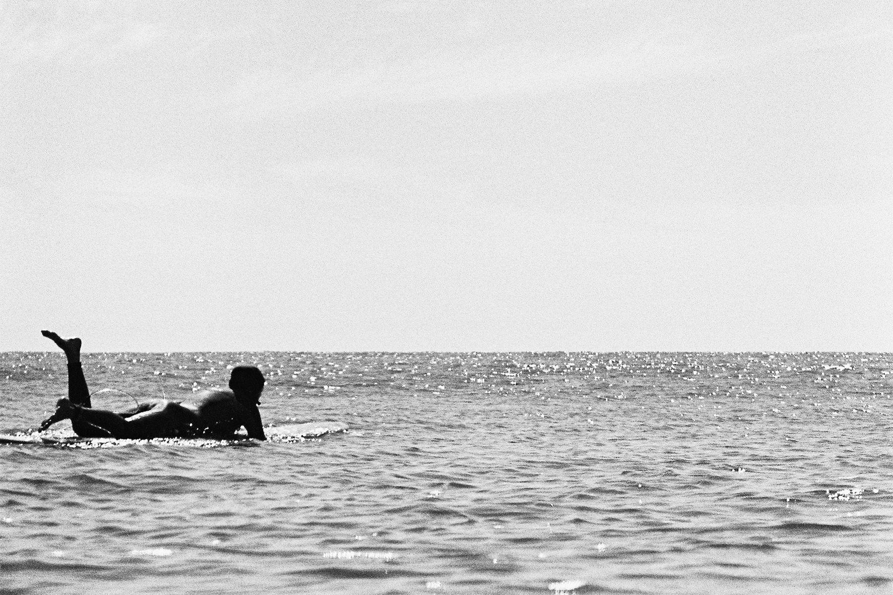 black and white photo of a longboarder lying on their board as they paddle back out, photographed on nikonos by grant musso